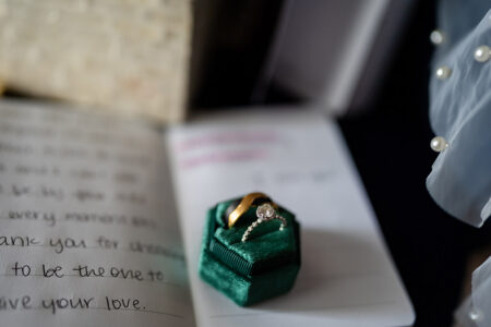 Diamond engagement ring and gold wedding band in green velvet box on handwritten letter, with fabric and pearls beside them.