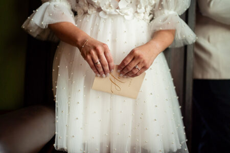 Torso and hands of someone in a pearl dress hold a gold-lettered card; another person in white hints at a New Orleans wedding.