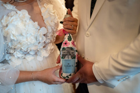 Couple in white holds floral-wrapped La Marca Prosecco, wedding rings and dress details visible—ideal Louisiana wedding inspiration.