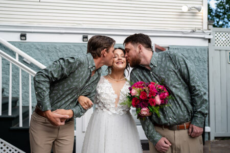 Bride in white dress smiles as two men in matching shirts kiss her cheeks at Louisiana wedding; she holds pink and red bouquet.