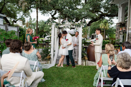 Bride and groom kiss under a floral arch at an outdoor Louisiana wedding, with guests, wedding party, and trees in the background.