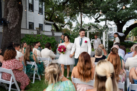Bride and groom hold hands at an outdoor New Orleans wedding, surrounded by clapping guests in a lush, garden setting.