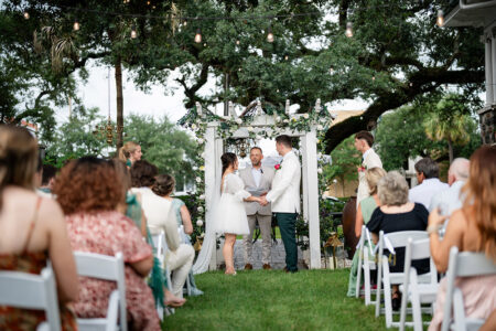 Bride and groom hold hands before an officiant under a decorated arch at a Louisiana outdoor wedding surrounded by guests.