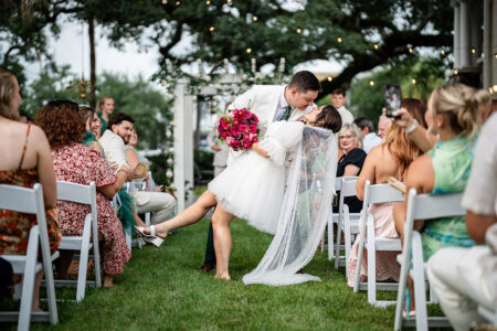 Groom in white suit dips and kisses bride in short dress as smiling guests take photos at outdoor New Orleans wedding.