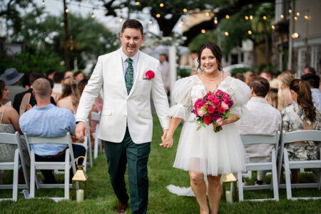 Bride and groom walk hand-in-hand down an outdoor Louisiana wedding aisle; she holds a pink bouquet, he wears green pants.