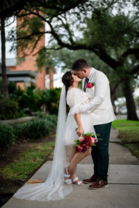 Bride and groom kiss on a lush, green sidewalk; she wears a short white dress and veil, he a white jacket—Gulf South wedding.