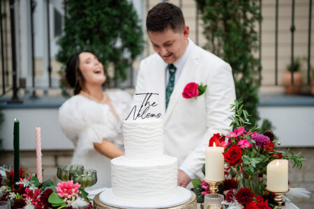 Bride and groom laughing by a white wedding cake with “The Nelsons” topper at an outdoor Louisiana reception.