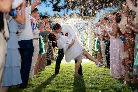 Couple kisses while dancing under string lights as guests blow bubbles at an outdoor Louisiana wedding.
