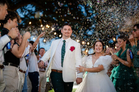 Newlyweds in white walk hand in hand through guests blowing bubbles under outdoor string lights at a Gulf South wedding.