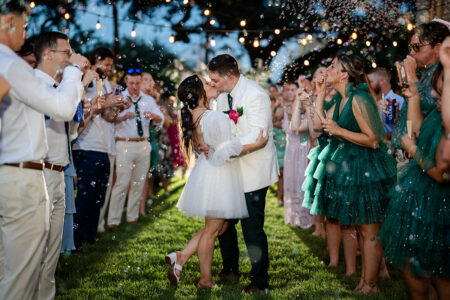 Bride and groom kiss under string lights as guests in festive attire cheer and blow bubbles at an outdoor wedding ceremony.