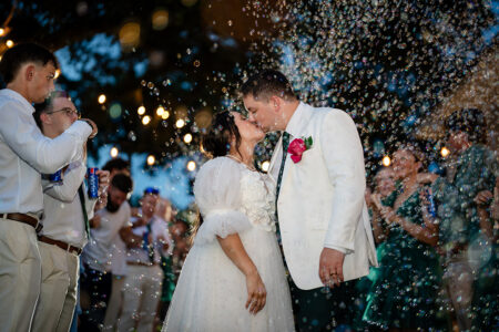 Bride and groom kiss under string lights at a Louisiana wedding, surrounded by bubbles, guests, and lush greenery.