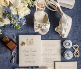Wedding essentials flat lay: white heels, bouquet, perfume, jewelry, blue ring box, garter, and invitations on blue surface.