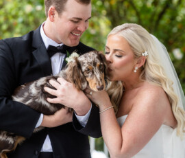 Bride and groom in wedding attire smile and kiss their brown and white dachshund outdoors at a joyful Louisiana wedding.