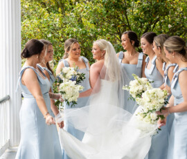 Bride in white gown with six bridesmaids in light blue dresses, all holding white bouquets on a porch at a Louisiana wedding.