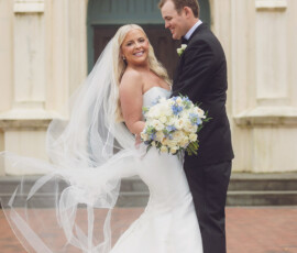 Bride in white gown and groom in black suit smile outside ornate New Orleans wedding venue.