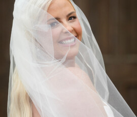Smiling bride with long blonde hair in a white veil and strapless dress looks over her shoulder against a dark background.