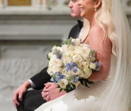 Smiling bride in white with blue bouquet sits by groom in black suit during New Orleans wedding in ornate venue.
