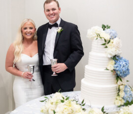 Bride and groom in formal wear smile with silver cups by a white wedding cake with blue flowers at their Louisiana celebration.