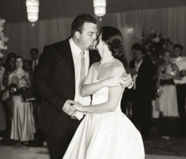 Newlywed couple sharing their first dance and a kiss on the reception dance floor surrounded by guests.