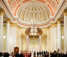 Elegant church wedding with domed ceiling, golden accents, and guests in pews facing the bride, groom, and wedding party at the altar.