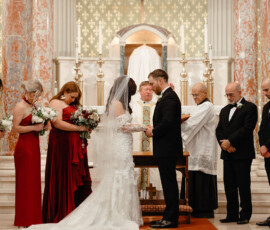 Bride and groom exchange rings at a downtown Mobile church altar, with bridesmaids, groomsmen, clergy, and crucifix in view.