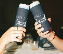 Two people toast cans in blue koozies with white line art at a Mobile Alabama wedding, drinks on a round table nearby.