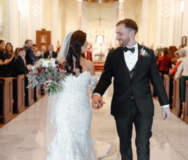 Bride and groom hold hands and smile as they walk down the aisle at their Mobile Alabama church wedding, guests watching.