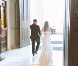 Bride in lace gown and groom in suit walk toward sunlight through open doors at an elegant downtown Mobile wedding.