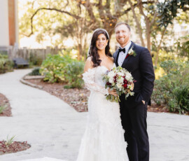 Bride in white gown and groom in black tuxedo smile on a sunny garden path at their downtown Mobile wedding.