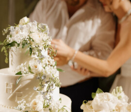 .Three-tier white wedding cake decorated with fresh white flowers while the couple cuts the cake in the background.