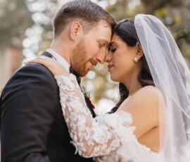 Bride and groom in wedding attire embrace outdoors, foreheads touching, eyes closed, with soft light and greenery in downtown Mobile.