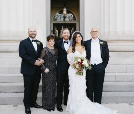 Bride and groom with family in formal attire pose outside a downtown Mobile building with columns, celebrating their wedding day.