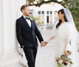 Bride in white dress and groom in black suit smile at each other by grand columns at a downtown Mobile wedding.