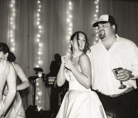 Bride in white dress dances and laughs with man in cap at Mobile, Alabama wedding, string lights glowing behind them.