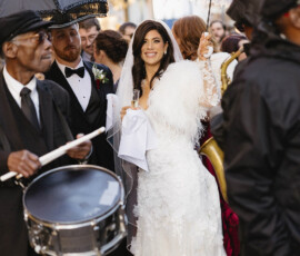 Bride in lace dress with white feathered umbrella walks joyfully with musicians in Cathedral Basilica Mobile wedding parade.