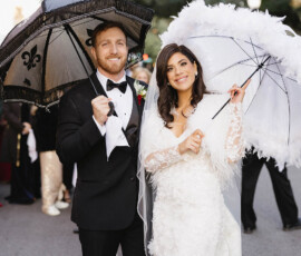 Bride and groom in formal attire smile with decorative umbrellas on a tree-lined downtown Mobile street, exuding wedding joy.