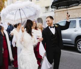 Bride and groom with decorated umbrellas parade down a downtown Mobile street, joined by smiling guests in red dresses.