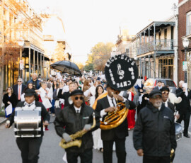 A brass band leads a festive parade in downtown Mobile, with people in formal wear and parasols, creating a joyful wedding vibe.