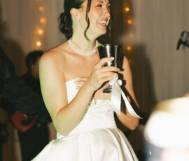 Bride in white dress and pearls holds silver cup before string lights at elegant Mobile cathedral wedding.
