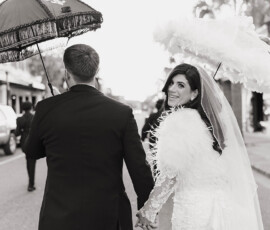 Bride and groom holding umbrellas walk hand in hand in downtown Mobile; bride smiles at the camera. Black and white photo.