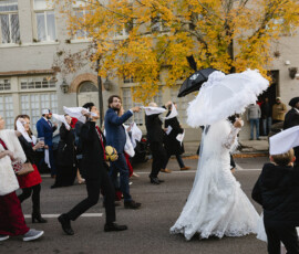 Bride in white gown leads downtown Mobile wedding parade with guests waving handkerchiefs; autumn trees and building behind.