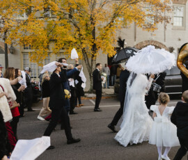 Bride and groom lead a joyful Mobile wedding parade with umbrellas, guests waving napkins, and a brass band on an autumn street.