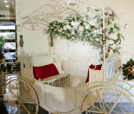Ornate white carriage seat with red cushions and floral decor, set indoors by a white brick wall for elegant weddings.