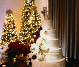 White tiered wedding cake with red and white flowers, bride and groom topper, gold candles, and Christmas trees in the background.