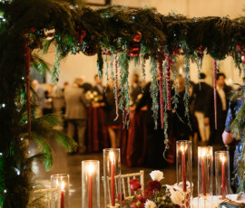 Elegant table with red candles, greenery, and flowers under an archway at a classic indoor wedding; guests mingle in background.