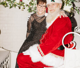 Smiling woman in sparkly black dress sits with Santa on white bench, festive flowers and greenery in downtown Mobile wedding setting.