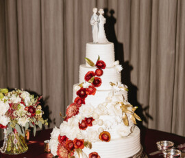 Three-tier white wedding cake with red and white flowers, bride and groom topper, on a red tablecloth with candles.