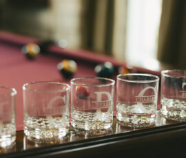 Five empty whiskey glasses on wood before a red pool table with scattered balls; sunlight streams through tall windows behind.