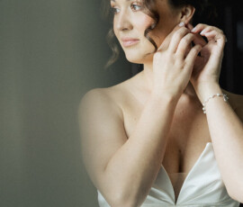 Bride in white strapless dress smiles softly indoors, putting on an earring before her Mobile Alabama wedding.