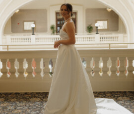 Bride in a strapless satin wedding gown posing beneath an ornate historic balcony inside the venue.